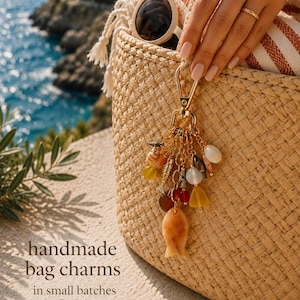 May include: A gold-toned metal bag charm featuring a collection of dangling elements including a carved fish pendant, various colourful glass beads, small seashells, and metallic charms, all attached to a lobster clasp hook on a woven straw bag.