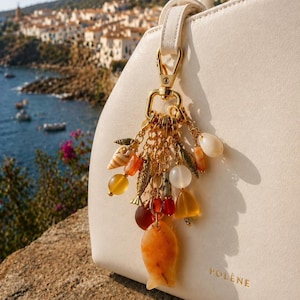 Puede incluir: Un charm dorado con varias cuentas y colgantes, incluyendo conchas marinas, peces y piedras preciosas, cuelga de un bolso blanco. El diseño del charm incorpora elementos naranjas, amarillos y blancos. El bolso tiene la marca "POLENE" impresa.