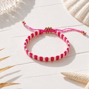 Könnte beinhalten: Ein rosa-weißes Perlenarmband mit Schiebeknotenverschluss. Das Armband hat abwechselnd rosa und weiße rechteckige Perlen. Die verstellbare Kordel ist rosa und hat goldfarbene Akzente. Das Armband wird auf einer weißen Oberfläche mit Muscheln präsentiert.