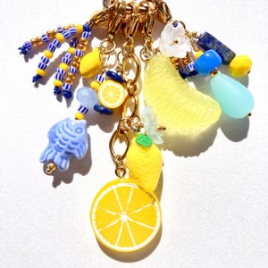 May include: A collection of gold-toned charms with fruit and sea-themed pendants. The charms include a lemon slice, a lemon, a fish, and a citrus fruit slice. The charms are accented with blue, yellow, and white beads.