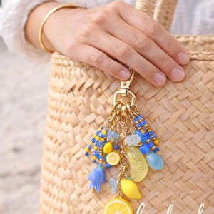 May include: A gold-toned keychain with a cluster of charms, including yellow lemon slices, blue beads, and a blue fish. The word "handmade" is visible. The keychain is attached to a woven straw bag.