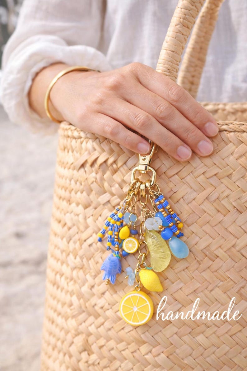 May include: A gold-toned keychain with a cluster of charms, including yellow lemon slices, blue beads, and a blue fish. The word "handmade" is visible. The keychain is attached to a woven straw bag.