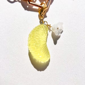 May include: A gold-colored charm with a yellow citrus fruit slice and a small white flower. The fruit slice is detailed with textured segments. The charm is attached to a gold chain.