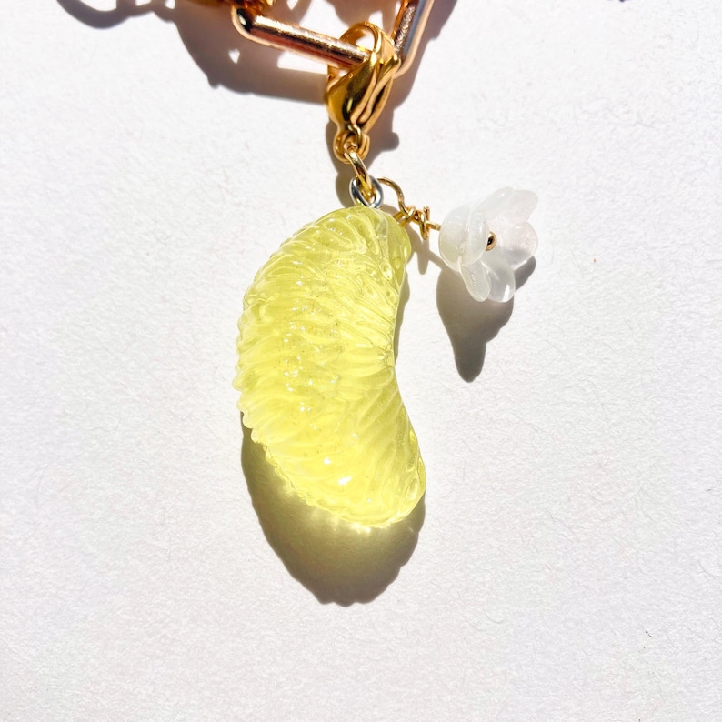 May include: A gold-colored charm with a yellow citrus fruit slice and a small white flower. The fruit slice is detailed with textured segments. The charm is attached to a gold chain.