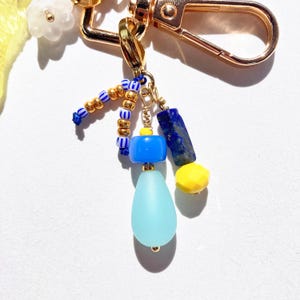 May include: A gold-colored keychain with various charms. The charms include blue, yellow, and gold beads, a blue teardrop-shaped bead, and a dark blue rectangular stone. The keychain has a lobster clasp.