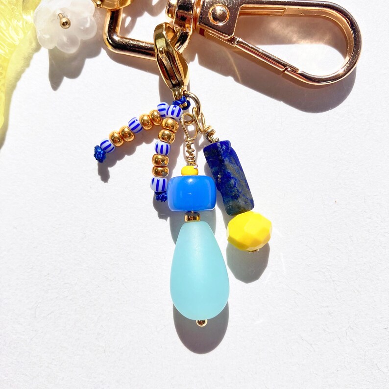 May include: A gold-colored keychain with various charms. The charms include blue, yellow, and gold beads, a blue teardrop-shaped bead, and a dark blue rectangular stone. The keychain has a lobster clasp.