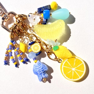 May include: A gold-toned keychain adorned with charms in a summery theme. The charms include yellow lemons, blue fish, and various blue, yellow, and white beads. A large yellow citrus slice is also present, creating a vibrant and playful accessory.