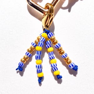 May include: Gold-toned metal charm with a beaded tassel. The tassel features strands of blue and white striped beads, yellow beads, and gold-colored beads. The charm is attached to a gold-colored clasp.