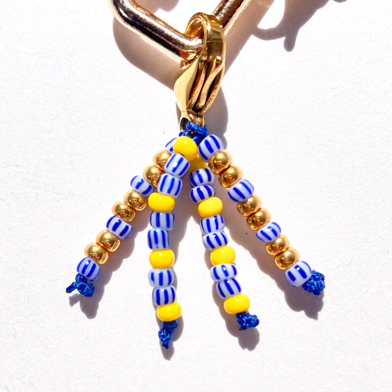 May include: Gold-toned metal charm with a beaded tassel. The tassel features strands of blue and white striped beads, yellow beads, and gold-colored beads. The charm is attached to a gold-colored clasp.