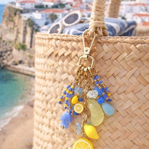Puede incluir: Un bolso tote de paja tejida con un cierre dorado y un dije decorativo. El dije presenta rodajas de limón amarillas, cuentas azules y amarillas y otros acentos coloridos. Gafas de sol y una toalla a rayas son visibles dentro de la bolsa.