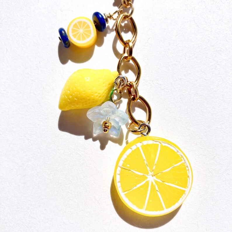 May include: A gold-colored chain with several charms. The charms include a lemon slice, a whole lemon, a blue flower, and blue and yellow beads. The lemon slice is yellow with white segments. The flower is light blue with a gold center.