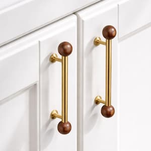 May include: Two cabinet pulls with a gold finish and dark brown wooden spheres at each end. The pulls are mounted on a white cabinet door. The hardware has a modern, minimalist design.