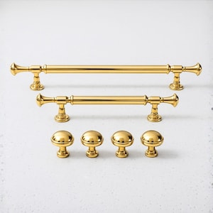 May include: A collection of gold-colored cabinet hardware, including two bar pulls and four round knobs. The bar pulls have a classic design with decorative end pieces. The knobs are a simple, rounded design. All pieces have a shiny, polished finish.