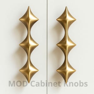 May include: Two brass-coloured cabinet pulls with a starburst design. Each pull has three diamond-shaped elements connected by a slender stem. The pulls are mounted on a white cabinet door. The text "MOD Cabinet Knobs" is printed below the pulls.
