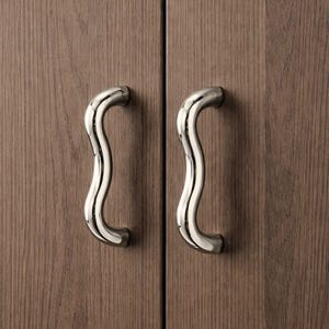 May include: Two polished chrome cabinet handles with a wavy design are mounted on a wooden cabinet. The handles are curved and have a smooth, reflective surface. The cabinet doors have a vertical wood grain pattern.