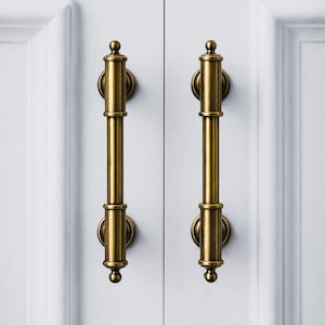 May include: Two antique brass cabinet pulls. Each pull has a cylindrical handle with decorative detailing and a rounded end. The pulls are mounted on a white cabinet door. The hardware has a warm, golden hue.