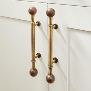 May include: Two cabinet pulls with a vintage aesthetic. Each pull features a dark brown wooden sphere at the top and bottom, connected by a textured, brass-colored rod. The pulls are mounted on a white cabinet door.