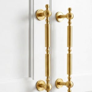 May include: Two gold-colored cabinet pulls with a vertical design. Each pull has a circular base, a central cylindrical section, and decorative spherical accents. The pulls are mounted on a white cabinet door.