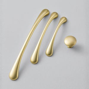 May include: A set of four gold-toned cabinet hardware pieces. The set includes three elongated pulls of varying sizes and a round knob. The hardware has a smooth, matte finish and a teardrop-shaped design.