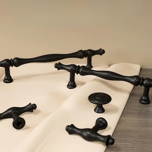 May include: A collection of matte black cabinet hardware including two long, curved drawer pulls with rounded ends, two smaller T-shaped pulls, and one circular knob. Each piece features a smooth, solid finish and a consistent design style.