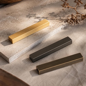 May include: Three rectangular metal cabinet pulls in gold, dark gray, and bronze tones. The pulls are arranged on a neutral-colored surface, with a wooden tray and dried plant sprig in the background. The pulls are a modern design.