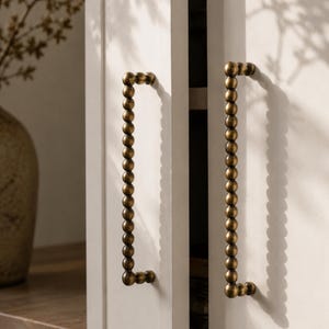 May include: Close-up of two white cabinet doors with brass-colored, beaded handles. The handles are rectangular with rounded corners and are attached to the doors. The cabinet doors are slightly ajar, revealing a glimpse of the interior.