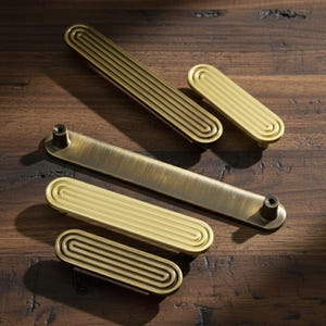 May include: A collection of four metal cabinet pulls in varying lengths. Three pulls feature a brushed gold finish with a repeating concentric oval pattern, while one longer pull has a smooth, brushed antique brass finish with two posts.