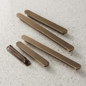 May include: A collection of bronze-colored cabinet pulls and handles. The hardware features a rectangular design with rounded ends. The pulls vary in size, with some having a single mounting post and others with two.