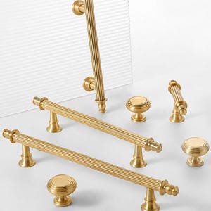 May include: A collection of brushed gold cabinet hardware including three long, fluted bar pulls with rounded ends and three matching round, tiered knobs. Each piece features a consistent metallic finish and vertical ribbed detailing.