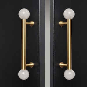 May include: Two cabinet door pulls. Each pull has a gold-colored, vertical bar with a white, marble-like sphere at each end. The pulls are mounted on a black cabinet door. The pulls have a classic, elegant design.