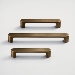 May include: Three bronze-colored cabinet pulls of varying lengths are arranged vertically against a white background. Each pull has a rectangular bar with rounded ends and a curved, rectangular base.