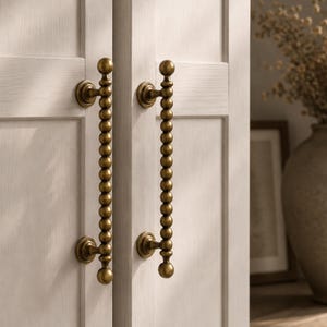 May include: Close-up of two cabinet doors with brass-coloured beaded door pulls. The pulls are vertically oriented and attached to the off-white cabinet doors. The hardware has a vintage aesthetic.