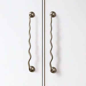 May include: Two vertical cabinet handles with a distinct wavy, serpentine silhouette. These metallic, silver-toned pulls feature rounded mounting bases at the top and bottom, securely attached to a smooth, solid white flat-panel cabinet surface.