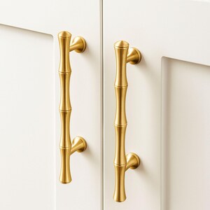 May include: Two gold-colored bamboo-style cabinet pulls mounted on a white cabinet door. The pulls are vertical, with a textured design mimicking bamboo stalks. The hardware is a warm, metallic tone, adding a touch of elegance.