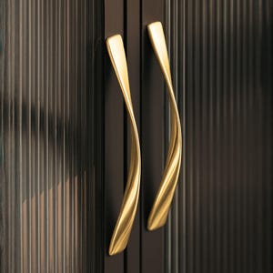 May include: Close-up of two gold-colored, curved cabinet handles. The handles have a smooth, polished finish and are mounted on a dark brown cabinet door with vertical ribbed detailing. The handles are slightly offset from each other.