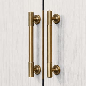 May include: Two gold-colored cabinet pulls with a vertical ribbed design. The pulls are mounted on a white wooden cabinet door. The hardware has a cylindrical shape with a circular base and a matching top.