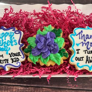 May include: Three decorated sugar cookies in a white box with pink shredded paper. The cookies are decorated with blue and white icing and the text "Happy Mother's Day from your favorite kid", a purple flower with green leaves, and "Thanks Mom I turned out awesome".