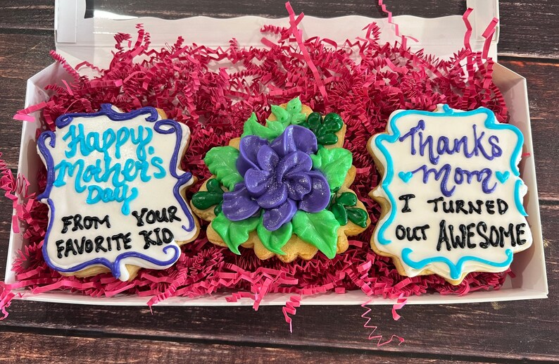 May include: Three decorated sugar cookies in a white box with pink shredded paper. The cookies are decorated with blue and white icing and the text "Happy Mother's Day from your favorite kid", a purple flower with green leaves, and "Thanks Mom I turned out awesome".