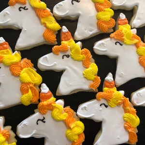 May include: White unicorn-shaped sugar cookies with orange, yellow, and white frosting. The cookies have a candy corn horn and a smiling face. The cookies are arranged on a dark surface.