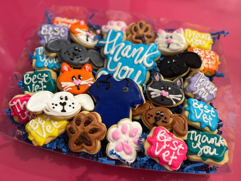 May include: A collection of decorated sugar cookies in a clear plastic container. The cookies feature various animal designs, including cats, dogs, and paw prints. Some cookies have the words "Thank You" and "Best Vet" written on them.