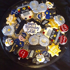 May include: A platter of decorated sugar cookies with a police theme. The cookies feature images of police cars, badges, handcuffs, guns, and the words "Happy Birthday Cash".