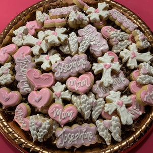 May include: A platter of decorated sugar cookies with pink and white icing. The cookies are shaped like hearts, crosses, wings, and have the names "Josephine" and "Mom" written on them.