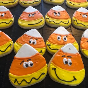 May include: A collection of Halloween-themed sugar cookies shaped and decorated to resemble candy corn. Each cookie features white, orange, and yellow icing, with black outlines and cartoon faces. The cookies are arranged on a dark wooden surface.