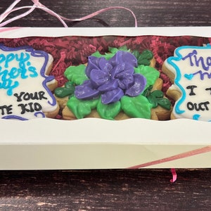 May include: A white gift box with a clear window showing three decorated cookies. The cookies are decorated with blue and purple icing and say "Happy Mother's Day from your favorite kid" and "Thanks Mom I turned out awesome".