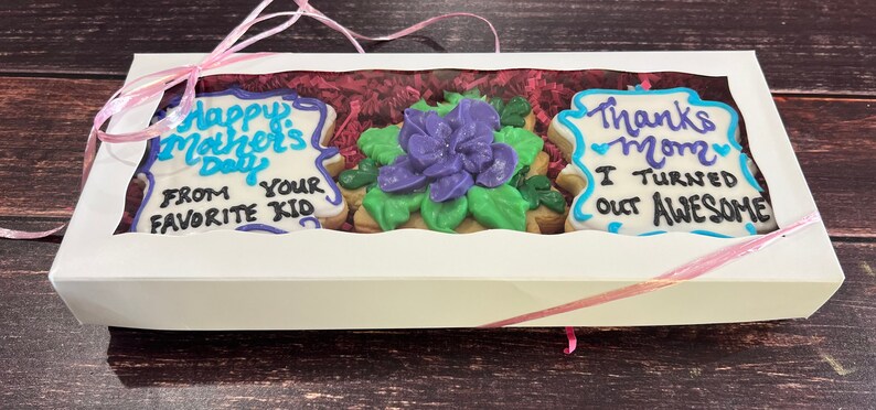 May include: A white gift box with a clear window showing three decorated cookies. The cookies are decorated with blue and purple icing and say "Happy Mother's Day from your favorite kid" and "Thanks Mom I turned out awesome".