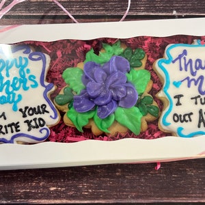 May include: A white gift box with a pink ribbon contains three decorated sugar cookies. The cookies are decorated with blue, purple, and green icing. The text on the cookies reads "Happy Mother's Day From Your Favorite Kid" and "Thanks Mom I Turned Out Awesome".