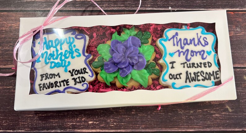 May include: A white gift box with a pink ribbon contains three decorated sugar cookies. The cookies are decorated with blue, purple, and green icing. The text on the cookies reads "Happy Mother's Day From Your Favorite Kid" and "Thanks Mom I Turned Out Awesome".