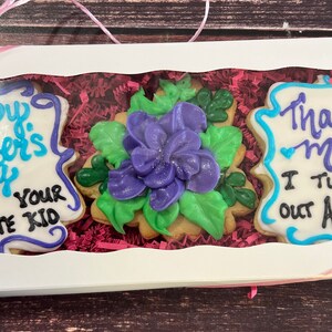 May include: A white gift box with a pink ribbon contains three decorated cookies. The cookies are decorated with blue and purple icing and feature the messages "Happy Mother's Day From Your Favorite Kid", "Thanks Mom I Turned Out Awesome", and a purple flower.