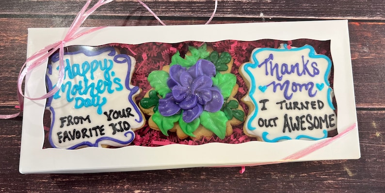 May include: A white gift box with a pink ribbon contains three decorated cookies. The cookies are decorated with blue and purple icing and feature the messages "Happy Mother's Day From Your Favorite Kid", "Thanks Mom I Turned Out Awesome", and a purple flower.