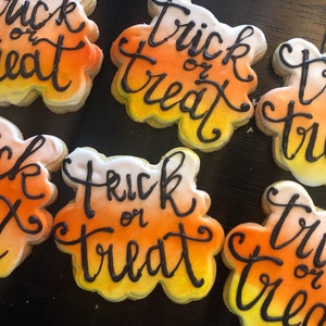 May include: Close-up of several Halloween-themed sugar cookies. Each cookie is shaped like a candy corn and decorated with orange, yellow, and white icing. The words "Trick or Treat" are written in black icing on each cookie. The cookies are arranged on a dark surface.